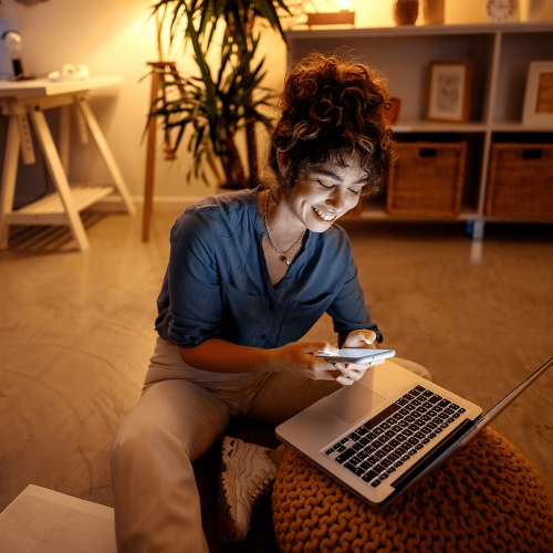 Smiling young woman sitting in front of laptop with mobile phone in her hands